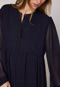 Svart klänning med ett mönster som har små blå och röda rutor, långa genomskinliga ärmar och en knäppt halsringning.