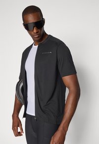 Schwarzes kurzärmliges Radsporttrikot mit Reißverschluss vorne, kombiniert mit einem weißen Mesh-Basisschicht und schwarzen gepolsterten Fahrradhosen.