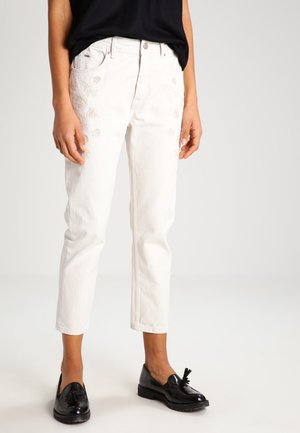 Osoba nosząca białe skrócone dżinsy z delikatnym kwiatowym haftem oraz czarne mokasyny z frędzlami, stojąca na jednolitym jasnym tle.