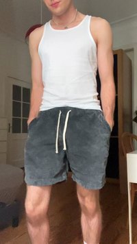 Pantaloni corti in velluto a coste grigi con coulisse, tasche anteriori e canotta bianca. Pavimento e mobili in legno visibili sullo sfondo.