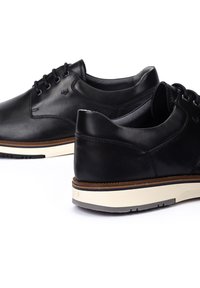 Zapatos de cuero negro con un acabado suave, punta redondeada y suela de goma blanca. Presentan cordones y acentos en contraste en marrón en la suela.
