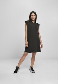 Zwarte mouwloze jurk met een relaxte pasvorm, zachte stof, ronde halslijn en subtiele naaddetails, gecombineerd met zwart-witte damborden sneakers.