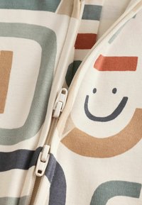 Färggädd tyg med abstrakta former och ett leende ansikte design, med en öppen dragkedja längs mitten. Mjuk textur syns.