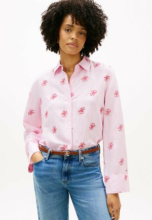 Kobieta o kręconych włosach, ubrana w jasnoróżową koszulę na guziki z czerwonymi nadrukami monogramów, niebieskie dżinsy i brązowy pasek.