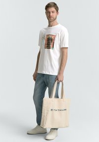 Hvid bomulds t-shirt med grafisk print, lyseblå jeans og beige tote bag med blåt logo. Hvide sneakers fuldender looket.