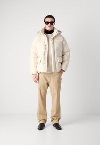 Hvid dunjakke med hætte, der har knaplukninger og forlommer. Sammen med beige bukser og sorte sko; modellen bærer mørke solbriller.