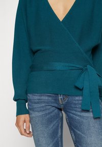 Kobieta ubrana w tealowy sweter z dzianiny typu wrap z bocznym wiązaniem oraz niebieskie, wyblakłe jeansy, z ręką swobodnie opartą na boku na jednolitym tle.