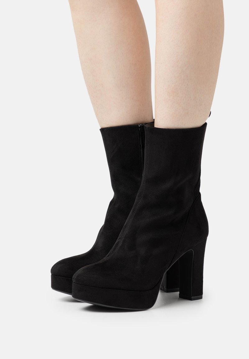 Bottines à plateforme en daim noir avec talons carrés portées sur jambes nues, présentées sur un fond clair uni.