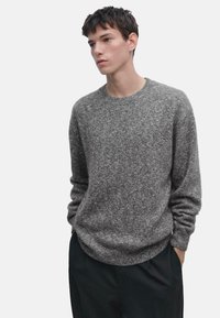 Maglione grigio lavorato a maglia con scollatura rotonda, polsini e orlo a coste. Indossato sopra pantaloni neri, con un aspetto morbido e testurizzato.