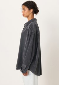 Ženska s kodrastimi lasmi, oblečena v preveliko temnosivo kordurovo srajco in svetlo modre jeans hlače, stoji v profilu na enobarvni podlagi.