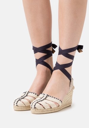 Espadrillas in tela beige con cuciture nere, tacco a zeppa e nastri incrociati neri intorno alle caviglie, suola in juta strutturata.