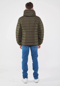 Giacca puffer verde oliva con design a trapunta diagonale, colletto alto e zip. Abbinata a jeans blu e sneakers bianchi.
