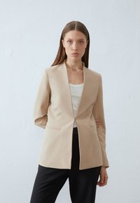 Blazer ajustado bege com colarinho recortado, fecho de botão único e dois bolsos laterais. Apresenta uma textura suave e uma silhueta estruturada.