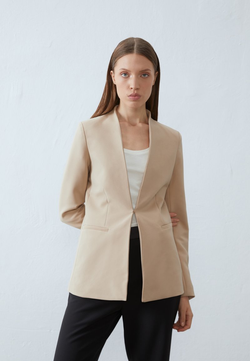 Blazer ajustado bege com colarinho recortado, fecho de botão único e dois bolsos laterais. Apresenta uma textura suave e uma silhueta estruturada.