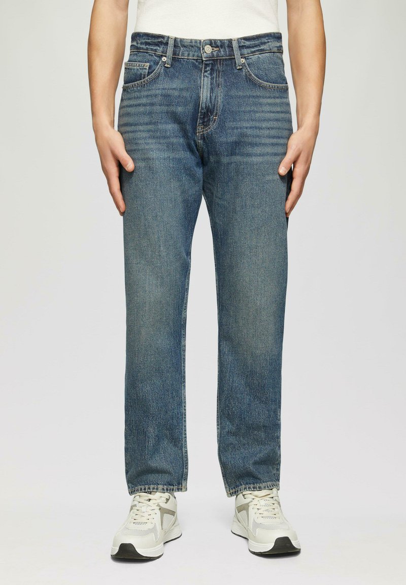 Herren Jeans aus blauem Denim, gerade geschnitten, mittlere Waschung, fünf Taschen, mit Metallknopf und Reißverschluss, kombiniert mit weißen und grauen Sneakers.
