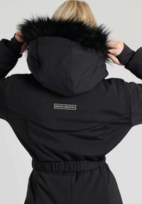 Schwarzer Kapuzen-Overall mit Kunstfellbesatz, tailliert und mit Logo-Patch. Verfügt über eine glatte Textur und funktionales Design.