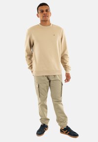 Felpa beige con colletto rotondo, abbinata a pantaloni cargo khaki chiaro con tasche con zip e sneakers blu navy con dettagli arancioni.