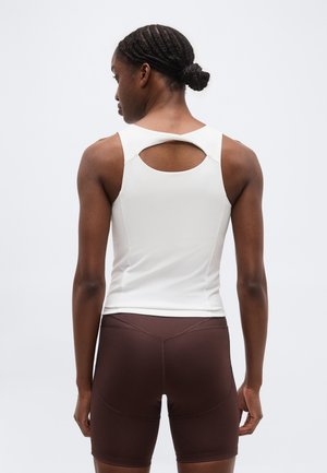 Γυναίκα με πλεγμένα μαλλιά σε χαμηλό σινιόν, φορώντας λευκό αμάνικο τοπ με άνοιγμα στην πλάτη και καφέ εφαρμοστά σορτς, στέκεται μπροστά σε ένα απλό φόντο.