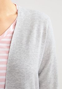 Svetle šedý cardigan vyrobený z měkké tkaniny, s otevřeným předním designem. Pod ním je částečně vidět růžové pruhované tričko s V výstřihem.
