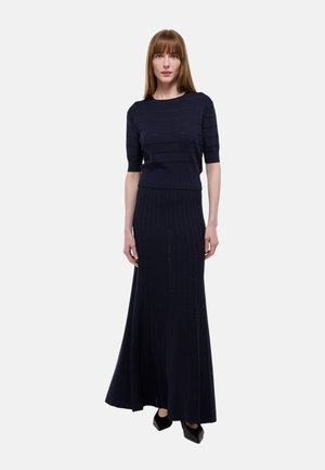 Femme portant un pull à manches courtes côtelé bleu marine et une jupe longue plissée assortie, avec des chaussures noires à bout pointu, debout devant un fond uni.