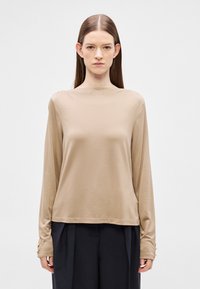 Beżowa bluzka z długim rękawem wykonana z gładkiej tkaniny, z wysokim dekoltem i guzikami na długich rękawach. Prosty, dopasowany krój.