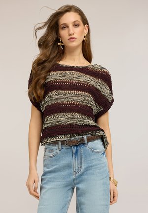 Młoda kobieta z długimi włosami, nosząca złote kolczyki, paskowany dzianinowy top, jasnoniebieskie dżinsy, brązowy pasek i złote bransoletki.