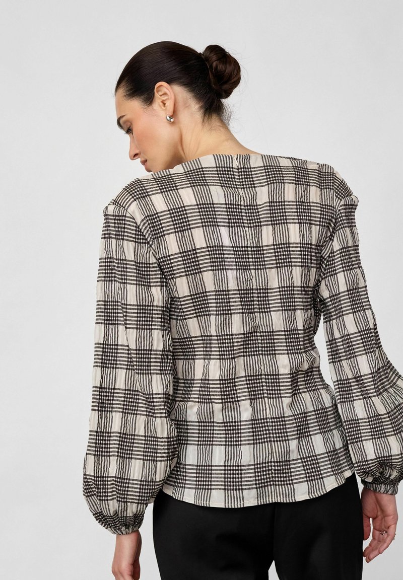 Γυναίκα με σκούρα μαλλιά σε κότσο, που φοράει μακρυμάνικο πουκάμισο σε ασπρόμαυρο καρό και μαύρο παντελόνι, με την πλάτη στραμμένη σε απλό φόντο.