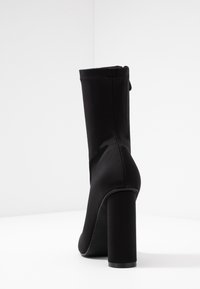 Bottine en tissu noir, talon bloc haut, texture lisse, fermeture éclair au dos, bout arrondi, détails cousus le long du bord supérieur.