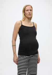 Nosečnica, oblečena v črno majico brez rokavov in črno-belo črtaste hlače, stoji pred enobarvnim belim ozadjem.