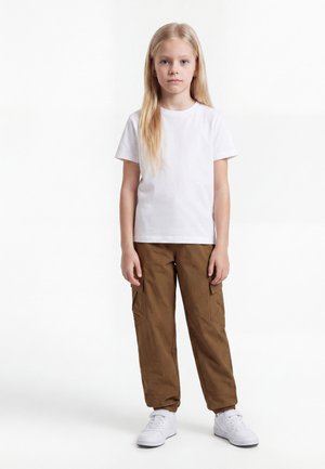 Lyshåret barn stående iført en ensfarvet hvid t-shirt, brune cargobukser og hvide sneakers, mod en hvid baggrund.