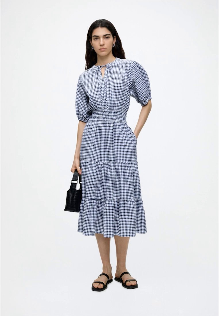 Femme portant une robe midi à carreaux bleus et blancs avec des manches bouffantes, tenant un petit sac à main noir et portant des sandales noires.