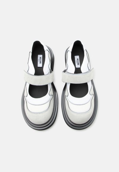 Paire de chaussures Mary Jane blanches et gris clair avec semelles noires et brides en Velcro, vue de dessus sur un fond clair.