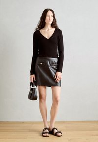 Haut noir côtelé à manches longues avec décolleté en V, associé à une jupe en cuir noire ajustée avec poches à rabat et boutons dorés, et des sandales noires.