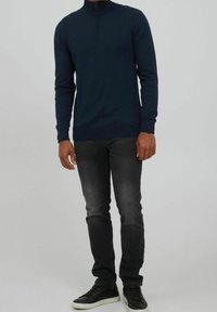 Granatowy sweter z wysokim kołnierzem i zamkiem, wykonany z miękkiego materiału. Połączony z czarnymi wąskimi dżinsami i czarnymi sneakersami.