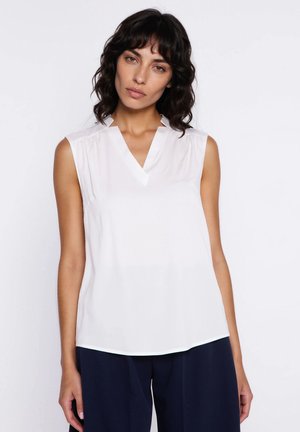 Kobieta o kręconych ciemnych włosach, ubrana w bezrękawową białą bluzkę z dekoltem w kształcie litery V oraz ciemnogranatowe spodnie na jednolitym tle.