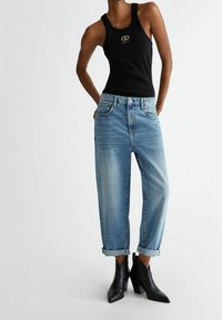 Nainen, jolla on yllään musta hihaton toppi, vaaleansiniset käännettävillä lahkeilla varustetut farkut ja mustat nilkkasaappaat, seisoo tasaisella taustalla.