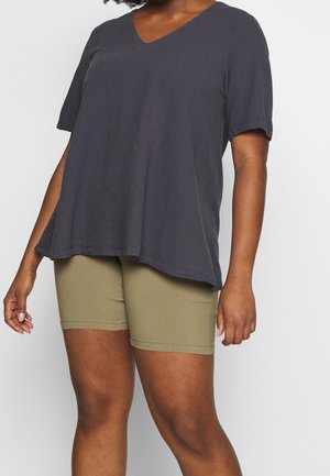 Kvinde iført løs, mørkegrå kortærmet V-hals top og tætsiddende olivengrøn cykelshorts, stående foran en ensfarvet baggrund.