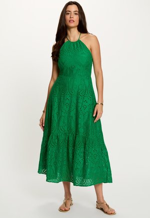 Femme debout portant une robe midi verte sans manches à œillets et des sandales dorées, aux longs cheveux foncés, sur un fond clair uni.