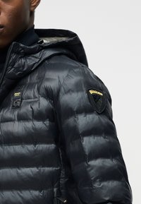 Μαύρο γυαλιστερό μπουφάν με επένδυση, με κουκούλα, που απεικονίζει μικρά κίτρινα λογότυπα στο στήθος και ένα έμβλημα σε σχήμα ασπίδας στο επάνω μέρος του μανικιού.