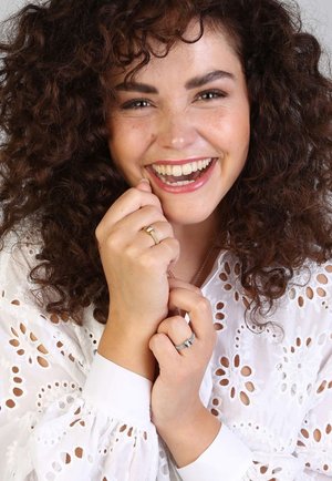 Model wearing a white, embroidered blouse with floral patterns. She has curly hair and showcases two rings, one gold and one silver.