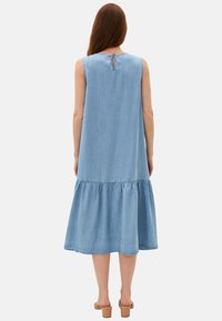 Vaaleansininen denimpitkäntyllinen mekko, jossa on kerrostettu helma ja takana avaus avaimenkolo. Liu'utuspinta, keskipitkä, rento siluetti.