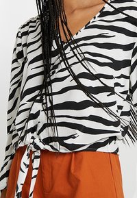 Blusa a righe zebrate in bianco e nero con scollatura a V e dettaglio a fiocco in vita, realizzata in un tessuto liscio, abbinata a pantaloni arancioni.