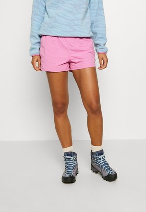 Person iført lyserøde shorts, lyseblå mønstret sweater, hvide sokker og grå vandrestøvler stående mod en ensfarvet hvid baggrund.