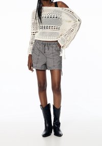Mujer vistiendo un top de crochet blanco de manga larga, pantalones cortos grises con cintura elástica y botas de vaquero negras hasta la mitad de la pantorrilla, de pie con una mano en el bolsillo.