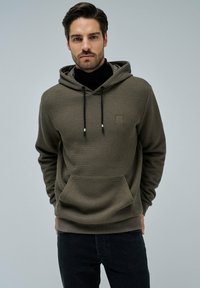 Felpa verde oliva con un design a maglia texturizzata, tasca frontale, cappuccio con coulisse e patch del marchio. Abbinata a jeans scuri.