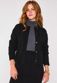 Cardigan nero a maglia con texture marmorizzata, dotato di colletto rotondo e chiusura a bottoni, abbinato a un dolcevita grigio.