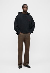 Homme debout les mains dans les poches, portant un sweat à capuche noir, un pantalon marron et des chaussures noires, sur un fond blanc uni.