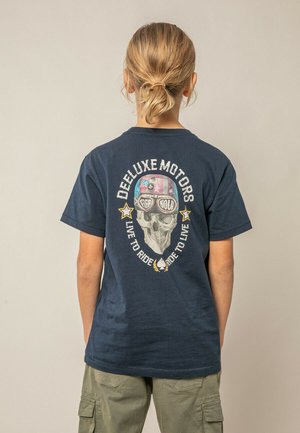 Personne aux cheveux blonds attachés en chignon porte un T-shirt bleu marine avec un graphique de crâne portant un casque et des lunettes, ainsi que le texte "Deeluxe Motors" au dos.