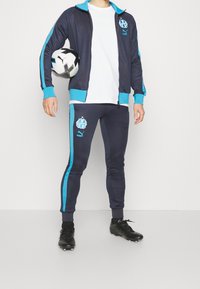 Marinblå träningsoverall med turkosa detaljer, bestående av en zip-up jacka och figurnära byxor. Inkluderar logotyper och en fotboll i personens hand.