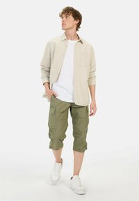 Beige Hemd mit Knöpfen über einem weißen T-Shirt, kombiniert mit olivgrünen Cargo-Shorts und weißen Sneakern mit braunen Akzenten.